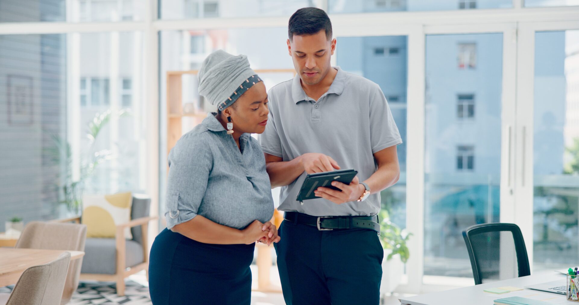 Pregnant woman in a gray head wrap and a man in a polo shirt review content together on a tablet in a modern office.